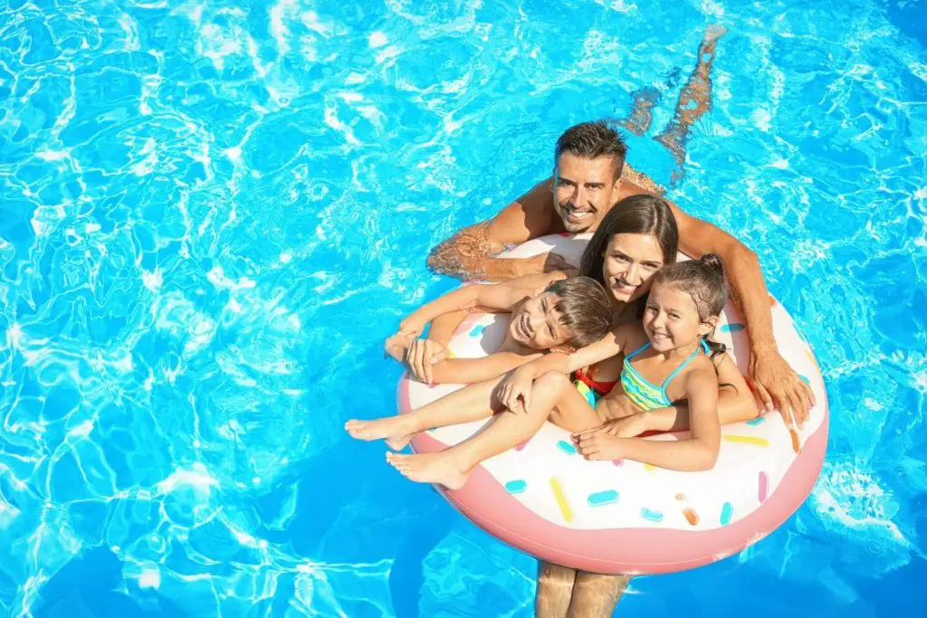 A smiling family of four enjoys a sunny day in a swimming pool, with the parents and two children sitting together on a donut-shaped inflatable float in clear blue water.