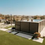 Four people are swimming in an above-ground pool in a modern backyard with artificial grass, potted plants, and lounge chairs. Two people are relaxing nearby under a clear blue sky.