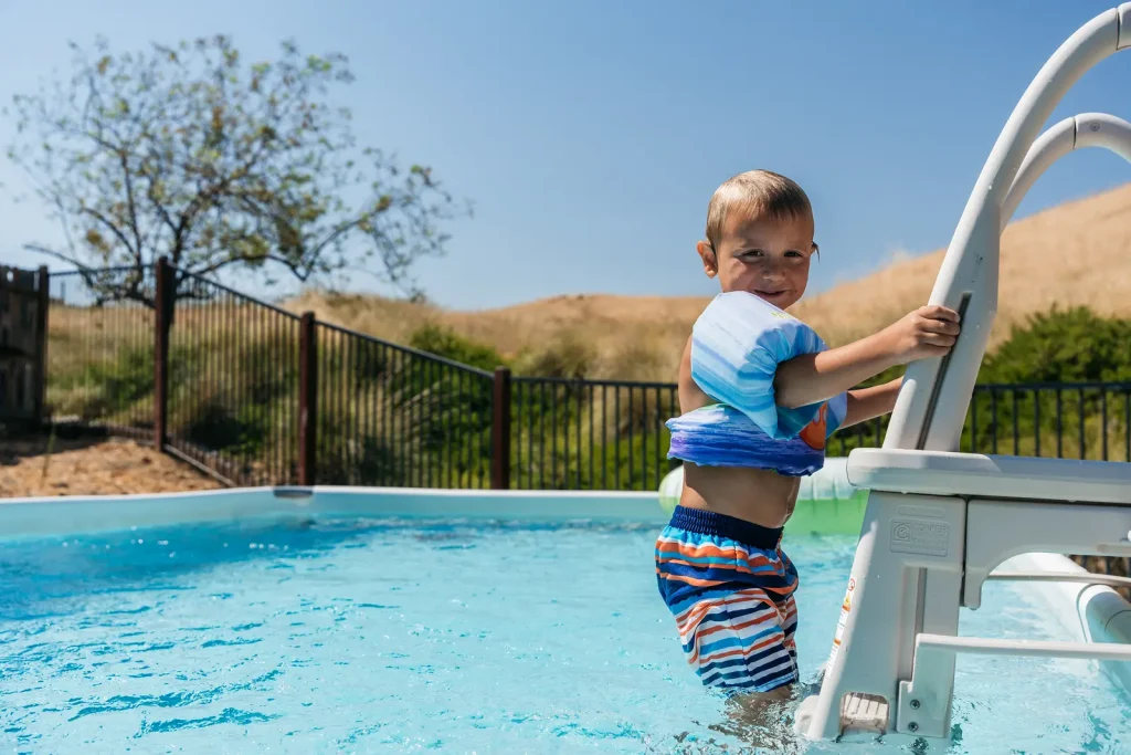 A young boy wearing a swim vest and striped swim trunks climbs a pool ladder in an outdoor above-ground pool on a sunny day, with a tree, fence, and grassy hills in the background.