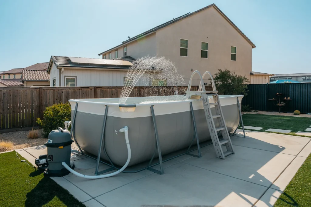 An above-ground backyard pool with a water fountain feature and ladder stands on a concrete patio. Houses, a wooden fence, and artificial grass are visible in the background on a sunny day.