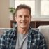 A middle-aged man with short brown hair sits on a couch, smiling warmly. He wears a blue plaid shirt over a gray t-shirt. The background shows a cozy living room with a window and some plants.