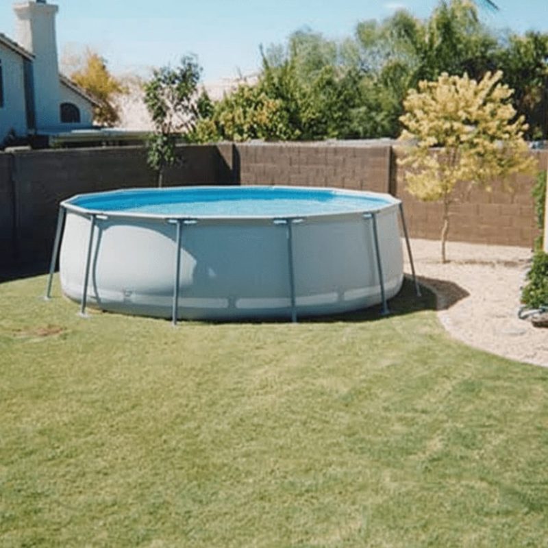 A round, above-ground pool sits on a grassy backyard lawn, surrounded by a brick wall and a few small trees and bushes, with houses visible in the background.